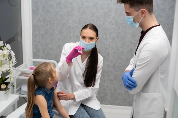 Una enfermera se está preparando para inyectar a una niña con una vacuna previamente preparada. El joven aprendiz intenta hablar con el niño para que no tenga miedo de la inyección. — Foto de Stock