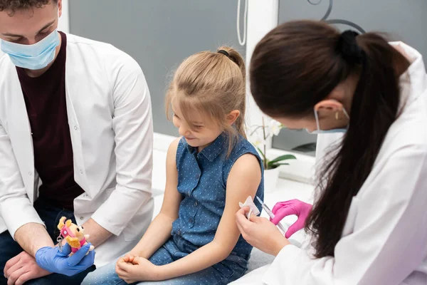 Una enfermera introduce una aguja en el brazo de las niñas con la nueva vacuna. Un médico senior anima a un pequeño paciente durante la vacunación. — Foto de Stock