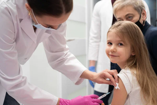 Una enfermera introduce una aguja en el brazo de las niñas con la nueva vacuna. La abuela saluda a su nieta durante la vacunación. El médico realiza la inyección necesaria en el brazo de los niños. Preventiva — Foto de Stock