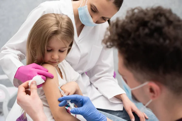 Doktor przygotowuje się do szczepienia małej dziewczynki. Młoda pielęgniarka siedzi obok dziecka, aby dziecko czuło się bezpiecznie. Szczepionka zapobiegawcza dla małych dzieci. — Zdjęcie stockowe