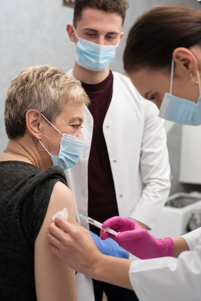 A nurse is making an vaccine to an elderly woman. The COVID vaccine19. A sterile doctors office in a private clinic. A young medical intern looks at how to properly inject.