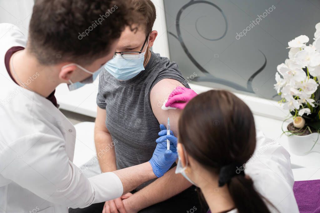 A young intern learns to inject a COVID19 vaccine into a young adult patient. A sterile doctors office in a private clinic. A young nurse teaches a medical intern how to properly inject.
