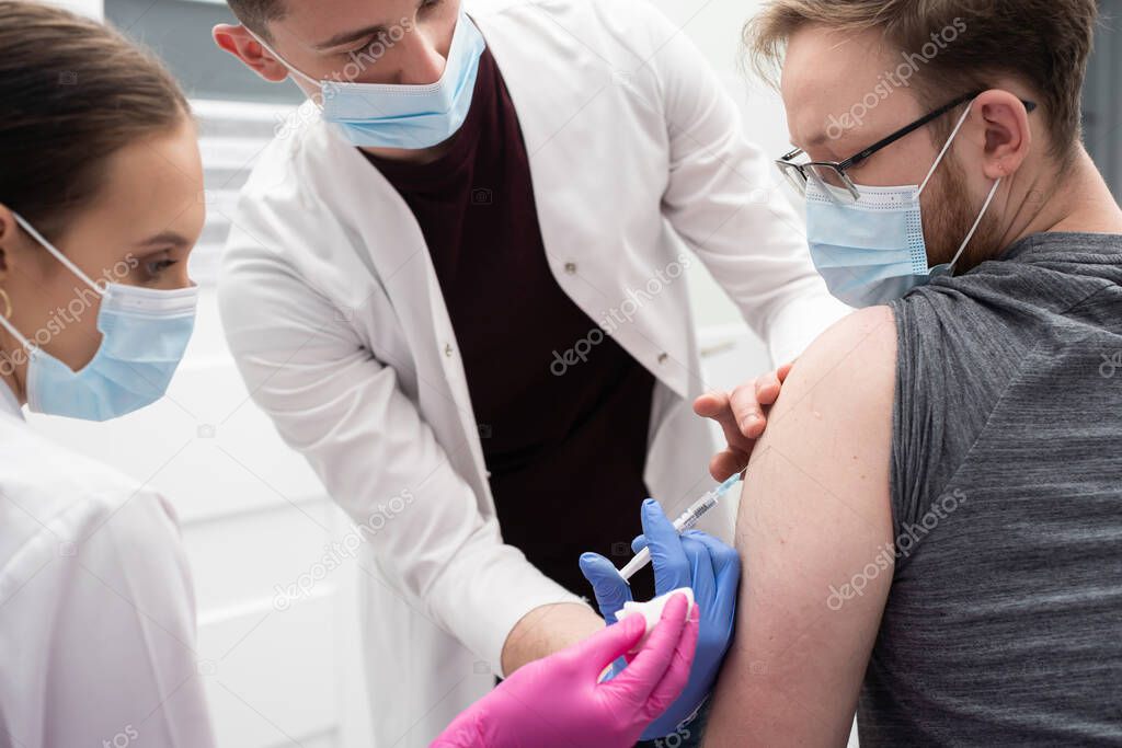 A young intern learns to inject a COVID19 vaccine into a young adult patient. A sterile doctors office in a private clinic. A young nurse teaches a medical intern how to properly inject.