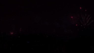 Ağaç tepelerindeki geniş çerçeve yeni yılın gelişini kutluyor. Periyodik olarak alevlerden ve diğer havai fişeklerden gelen parlak ışıklar koyu arkaplana çarpıyor.