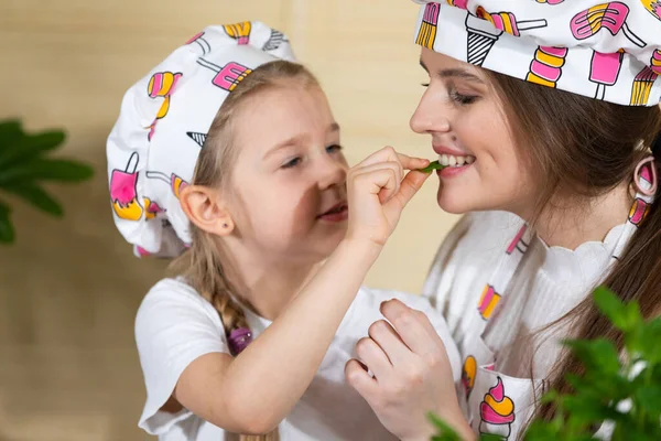 Ao decorar a pizza caseira com ervas, minha filha decidiu dar uma folha verde fresca para a mana para tentar. Mãe e filha momentos juntos na cozinha da casa. — Fotografia de Stock