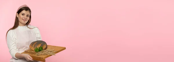 Panoramic frame. A young attractive saleswoman from a bakery shop holds a round loaf of bread sprinkled with poppy seeds and a pinch of fresh green herb leaves and rye ears on a wooden tray. The whole — Stock Photo, Image