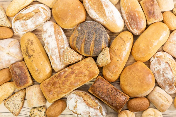 Many kinds of bread and buns randomly placed next to each other on a wooden countertop as decoration of a traditional bakery with a multi-generational experience. View from above. — Stock Photo, Image