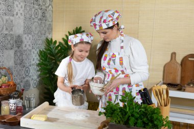Anne ve kız hamur yoğurmak için malzemeleri hazırlıyorlar ve buğday ununu süzgeçten geçirip mutfak tahtasına döküyorlar..