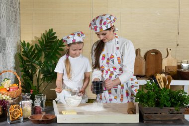 Anne ve kız hamur yoğurmak için malzemeleri hazırlıyorlar ve buğday ununu süzgeçten geçirip mutfak tahtasına döküyorlar..