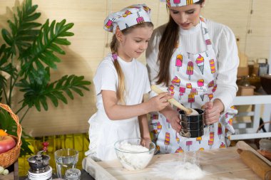 Anne ve kız hamur yoğurmak için malzemeleri hazırlıyorlar ve buğday ununu süzgeçten geçirip mutfak tahtasına döküyorlar..