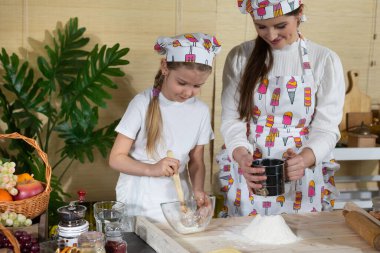 Anne ve kız hamur yoğurmak için malzemeleri hazırlıyorlar ve buğday ununu süzgeçten geçirip mutfak tahtasına döküyorlar..