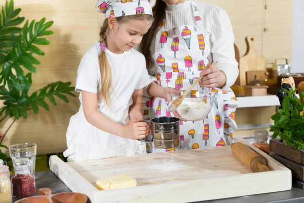 Anne ve kız hamur yoğurmak için malzemeleri hazırlıyorlar ve buğday ununu süzgeçten geçirip mutfak tahtasına döküyorlar..