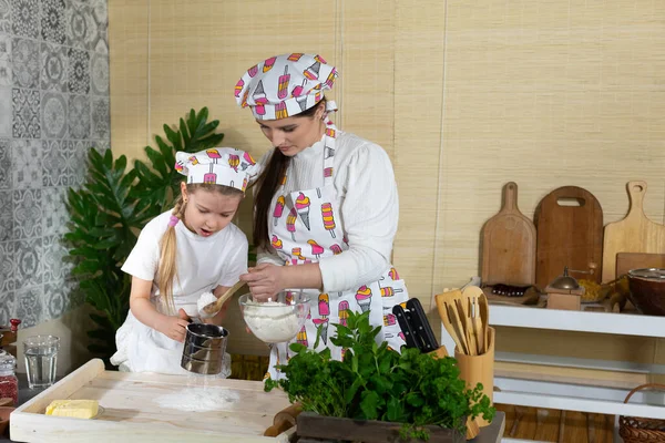 Anne ve kız hamur yoğurmak için malzemeleri hazırlıyorlar ve buğday ununu süzgeçten geçirip mutfak tahtasına döküyorlar..