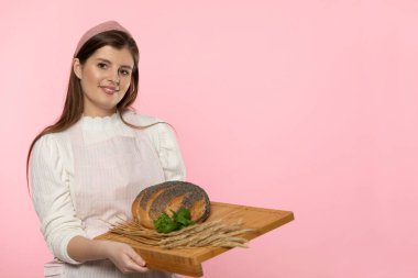 Bir fırından genç ve çekici bir satıcı elinde haşhaş tohumları serpiştirilmiş yuvarlak bir somun ekmek ve bir tutam taze yeşil ot yaprağı ve ahşap tepside çavdar kulağı tutuyor. Üniformalı olan.