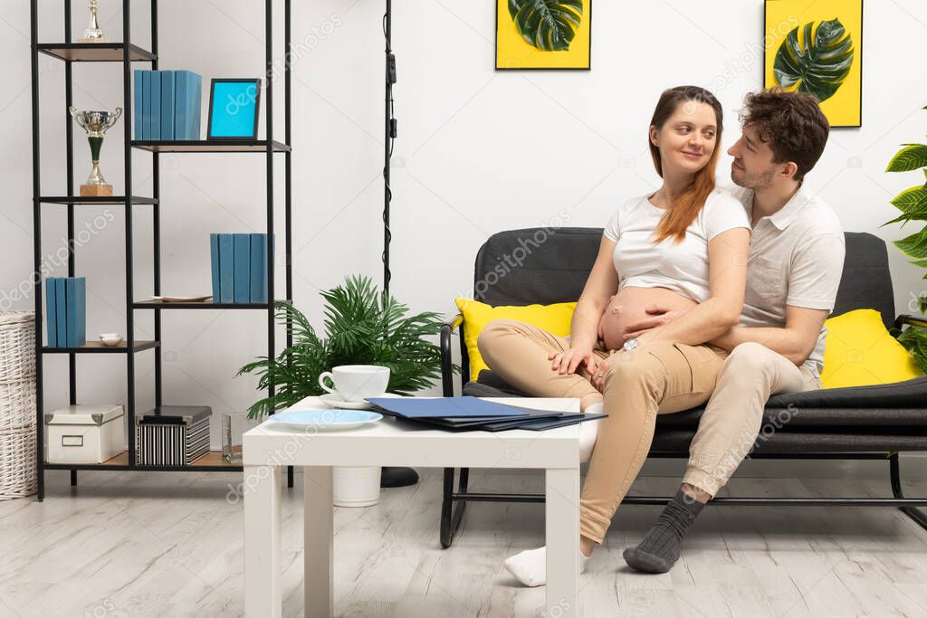 At home, a married couple sits on the sofa and admire her big belly because the woman is heavily pregnant. A bright living room in a modern style with a preferable space.