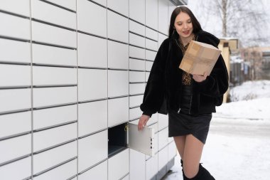Uzun boylu bir kız elinde bir paketle paket dolabının önünde duruyor. Kışın paketleri otomatik olarak al ve gönder.