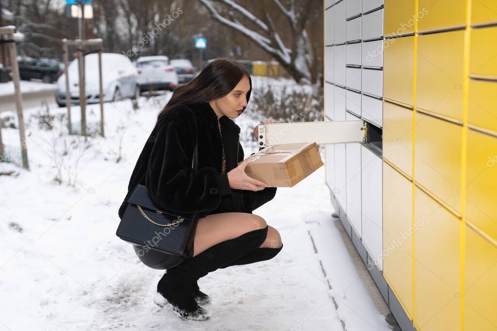 The student crouched at the parcel locker and holds a gray package in her hand. Receive and send parcels automatically in winter.