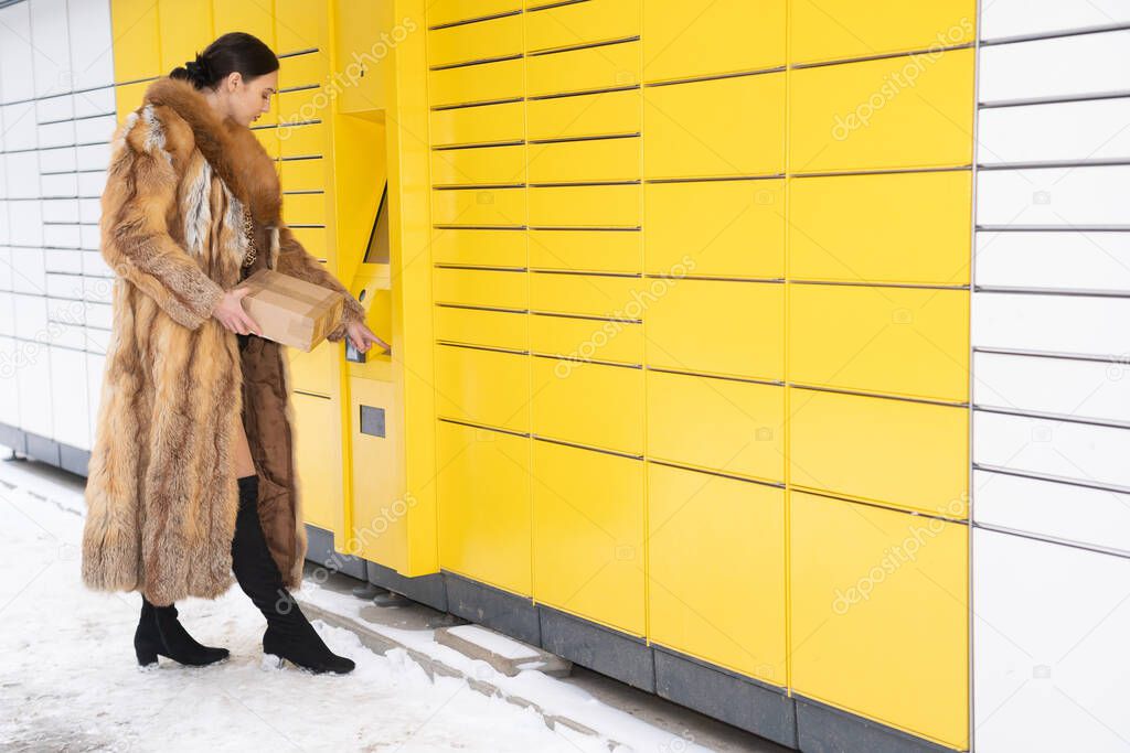A woman in a fur coat types the code on the numeric keypad of a parcel locker. Receive and send parcels automatically in winter.