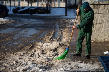 Elle çalışan bir işçi, ıslak bir kaldırımdan bir süpürgeyle kar kalıntılarını temizler.