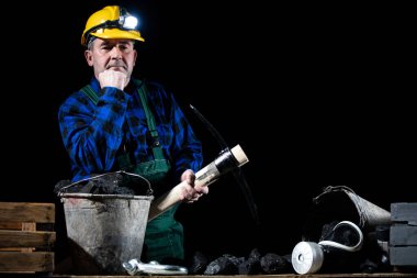 Elinde kazma olan bir madenci, siyah arka planda siyah kömür dolu bir kovanın yanında duruyor.