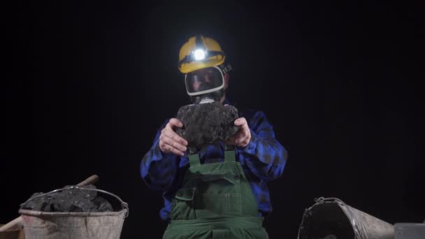 Un mineur portant un masque anti-poussière et un casque avec une lampe de poche tient une boule de charbon noir dans ses mains — Video