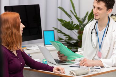 Bir doktor yüzünde gülümseme olan kızıl saçlı bir hastayla konuşur. Doktor muayenehanesinde yetişkin bir kadın.
