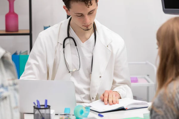 A doctor checks a book calendar for the date of the next follow-up appointment for a young teenage girl. The doctors office