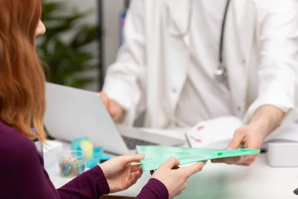 Una paciente con el pelo rojo le da los registros médicos a su médico de familia. Oficina de médicos. Vista de primer plano —  Fotos de Stock