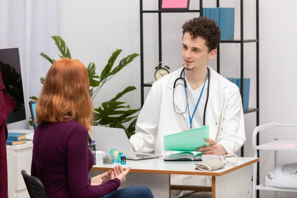 Un médico habla con un paciente pelirrojo con una sonrisa en la cara. Una mujer adulta en un consultorio médico —  Fotos de Stock