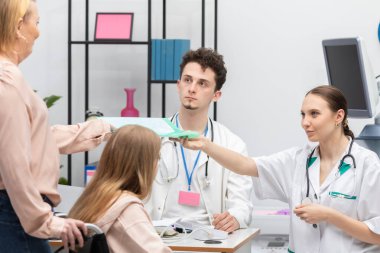 Bir hemşire, kızlarına test sonuçlarını annesine verir. Tekerlekli sandalyede bir genç ve annesi muayenehanede bekliyor..