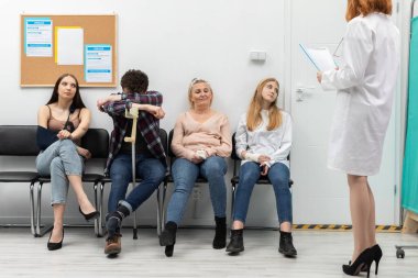 Ofisinin önünde duran doktor bir sonraki kişiyi okur ve onu check-up için içeri davet eder. Farklı yaşlarda insanlar sandalyede oturuyor.