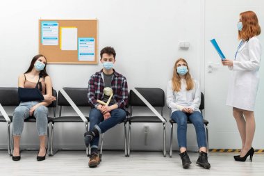 Doktor sıradakini yüksek sesle okur. Salgın sırasında herkes koruyucu maske takmak zorundadır.