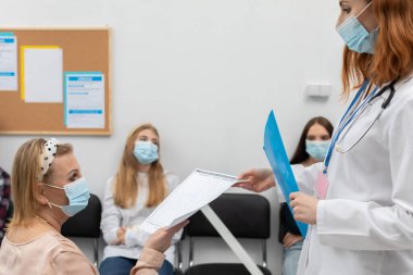 Kızıl saçlı bir doktor doktorunun muayenehanesinin önünde duruyor ve COVID-19 ile ilgili broşürleri okuyor. Ağzının ve burnunun üzerinde koruyucu bir maske..