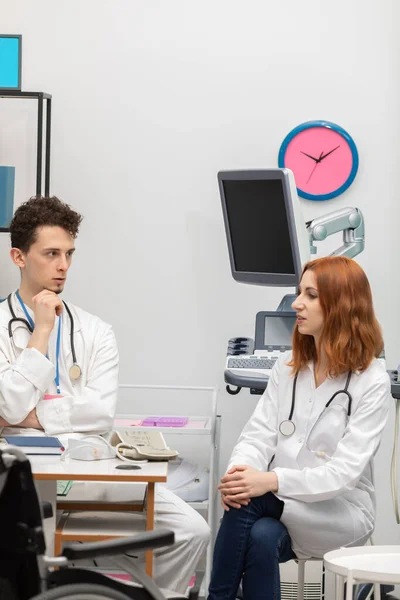 Un joven doctor habla con un interno pelirrojo sobre su próximo paciente. GP. Oficina de médicos —  Fotos de Stock