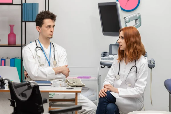 Un joven doctor habla con un interno pelirrojo sobre su próximo paciente. GP. Oficina de médicos —  Fotos de Stock