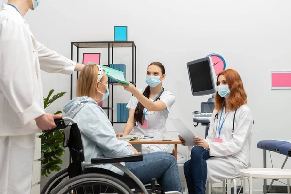 Un médico le da a un paciente resultados de pruebas a un enfermero practicante. Una mujer de 50 años en silla de ruedas en el consultorio médico —  Fotos de Stock