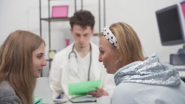 De dochter en moeder kletsen graag in het kantoor terwijl de dokter de medische dossiers opstelt. — Stockvideo