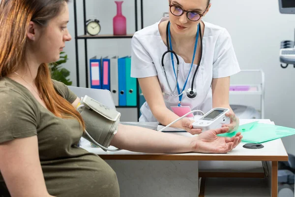 A lady doctor checks the result after testing a pregnant patient with a blood pressure monitor. A married couple in a gynecologists office.