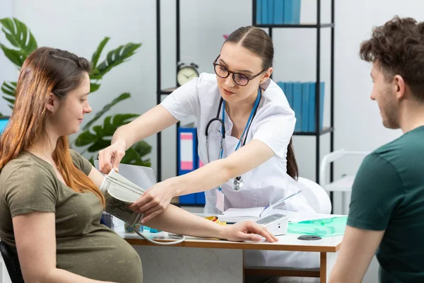 Un ginecólogo coloca un dispositivo de medición de la presión arterial en el brazo de una mujer embarazada. Una pareja casada en una oficina de ginecólogos. — Foto de Stock