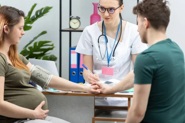 Eine Ärztin notiert das Ergebnis eines Blutdruckmessgeräts für schwangere Patientinnen in das Diagramm. Ein Ehepaar in einem Frauenarztbüro. — Stockfoto