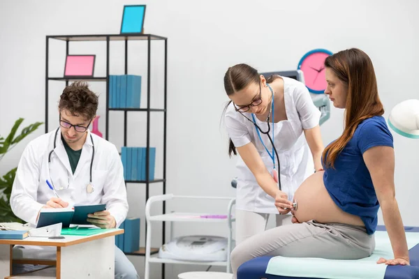 Ihr Arzt untersucht den Bauch einer schwangeren Patientin mit einem Stethoskop. Der behandelnde Arzt ergänzt den Terminkalender. — Stockfoto