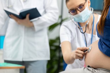 Koruyucu maske takan bir bayan doktor hamile bir hastanın steteskopuyla karın bölgesini inceliyor. Salgın sırasında.