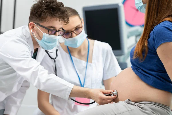 Un médico revisa el abdomen de una paciente embarazada con un estetoscopio. Máscaras protectoras colocadas sobre la boca y la nariz durante una pandemia. Vista de primer plano. — Foto de Stock