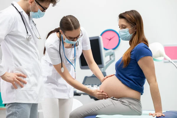 Eine Krankenschwester untersucht unter Anleitung eines Arztes den Bauch einer schwangeren Patientin mit ihren Händen. Schutzmasken bei Pandemie über Mund und Nase gelegt. — Stockfoto