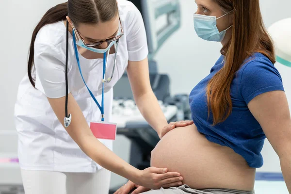 Eine Ärztin untersucht mit ihren Händen den Bauch einer schwangeren Patientin. Schutzmasken über Mund und Nase während einer Pandemie. — Stockfoto
