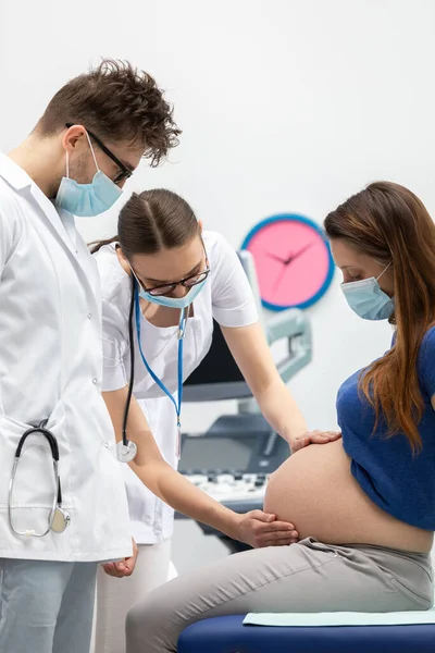 Eine Krankenschwester untersucht unter Anleitung eines Arztes den Bauch einer schwangeren Patientin mit ihren Händen. Schutzmasken bei Pandemie über Mund und Nase gelegt. — Stockfoto