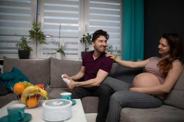 Ein Ehemann massiert seine Frau mit den Füßen und sie sitzen zusammen auf dem Sofa im Wohnzimmer. Schwangere. — Stockfoto
