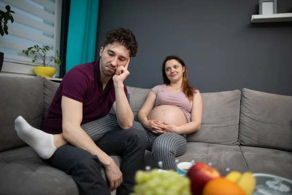 Ein Ehepaar sitzt zu Hause auf dem Sofa und der Ehemann macht sich Gedanken darüber, wie es nach der Geburt sein wird. Schwangere. — Stockfoto