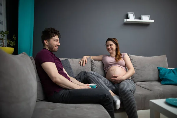 Un marido se sienta con una taza de café en el sofá y sonríe a su esposa embarazada. Una mujer con un embarazo avanzado. — Foto de Stock