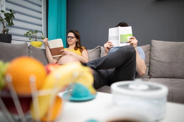 A married couple is reading a book on parenting and the husband is particularly engrossed and puts the book under his nose. A woman in advanced pregnancy. — Stock Photo, Image